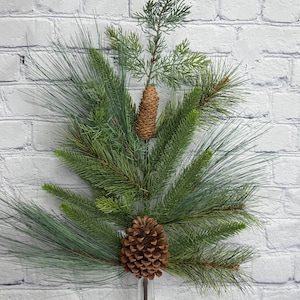 May include: A decorative arrangement of evergreen branches and pinecones in a clear glass vase. The arrangement features long, needle-like pine needles and smaller, textured pinecones, creating a natural, rustic aesthetic. The background is a white brick wall.
