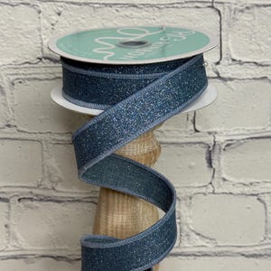 May include: A spool of blue glitter ribbon with a light blue edge. The ribbon is wrapped around a wooden spool. The ribbon is a decorative craft supply, perfect for gift wrapping, crafting, or adding a touch of sparkle to any project.
