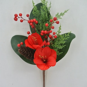 May include: A decorative floral stem featuring two vibrant red amaryllis blooms, complemented by green foliage, red berries, and a small pine cone. The stem is brown. This artificial arrangement is suitable for holiday or seasonal decor.