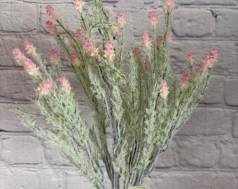 May include: A bouquet of artificial pink and white flowers with green stems. The flowers are arranged in a clear glass vase. The vase is sitting on a white brick wall. The flowers have a frosted look. A green tag with the text "Regency Home Decor" is attached to the vase.