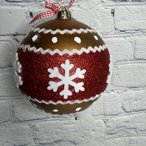 May include: A round Christmas ornament with a bronze-colored top and a red glittery band. The ornament features white snowflake and dot designs, with a white wavy trim. It hangs from a red and white striped string and a red and white checkered bow.