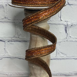 May include: A spool of decorative ribbon featuring a central band of shimmering orange glitter flanked by metallic bronze edges. The ribbon is wound around a circular spool and displays a textured, sparkling finish with a distinct woven border.