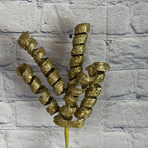 May include: A decorative gold glitter spiral stem. The stem has multiple curled spirals with a glittery texture. The stem is green and is in a clear vase. The background is a white brick wall.