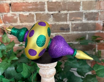 May include: Two decorative ornaments. One is gold with purple and green polka dots, the other is purple with a green base and yellow tip. Both are glittery and have gold hanging loops. They are displayed on a wooden stand.
