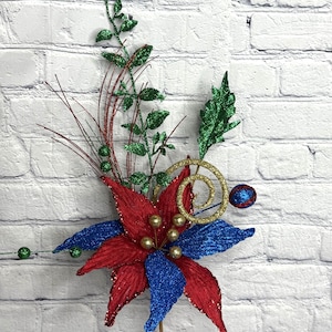 May include: A festive floral arrangement featuring a large red and blue poinsettia with gold accents. Green leaves and berries, along with a gold spiral, add to the holiday theme. The arrangement is on a clear stem.