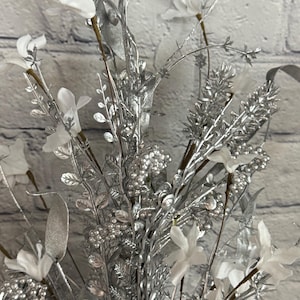 K&ouml;nnte beinhalten: Nahaufnahme eines dekorativen Blumenarrangements mit silberfarbenen Zweigen, Bl&auml;ttern und Beeren. Wei&szlig;e Stoffblumen sind in der Anordnung verstreut. Der Hintergrund ist eine hellgraue Backsteinmauer.