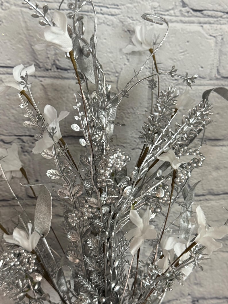 K&ouml;nnte beinhalten: Nahaufnahme eines dekorativen Blumenarrangements mit silberfarbenen Zweigen, Bl&auml;ttern und Beeren. Wei&szlig;e Stoffblumen sind in der Anordnung verstreut. Der Hintergrund ist eine hellgraue Backsteinmauer.