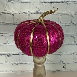 Puede incluir: Una calabaza decorativa fucsia con lentejuelas, detalles dorados y un tallo dorado. La calabaza se exhibe sobre un pedestal de madera de color claro. El fondo es una pared de ladrillos blancos.