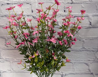 Puede incluir: Un ramo de flores artificiales rosas con hojas verdes en un jarrón de vidrio transparente sobre un fondo de pared de ladrillo blanco.