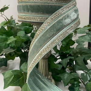 May include: A spool of sage green velvet ribbon with gold trim. The ribbon is displayed on a decorative stand, with a lush green ivy background. The ribbon is approximately 2 inches wide.