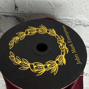 Op de afbeelding: Een ronde zwarte spoel met donkerbordeauxrood lint. De bovenkant van de spoel is voorzien van een gele lauwerkransafbeelding en de tekst John Mark Enterprises in het geel gedrukt. Het lint is om de spoel gewikkeld met een gladde, satijnachtige afwerking.