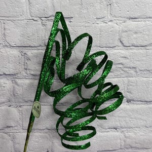 May include: A decorative green glitter ribbon stem with a tag. The stem is a dark green color and has a series of spiraling loops. The background is a white brick wall.