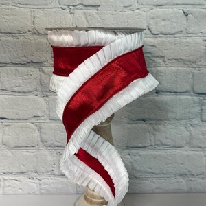 May include: A spool of red and white decorative ribbon. The ribbon features a wide, shiny red satin stripe bordered by a ruffled white fabric. The ribbon is displayed on a wooden spool, against a white brick wall.