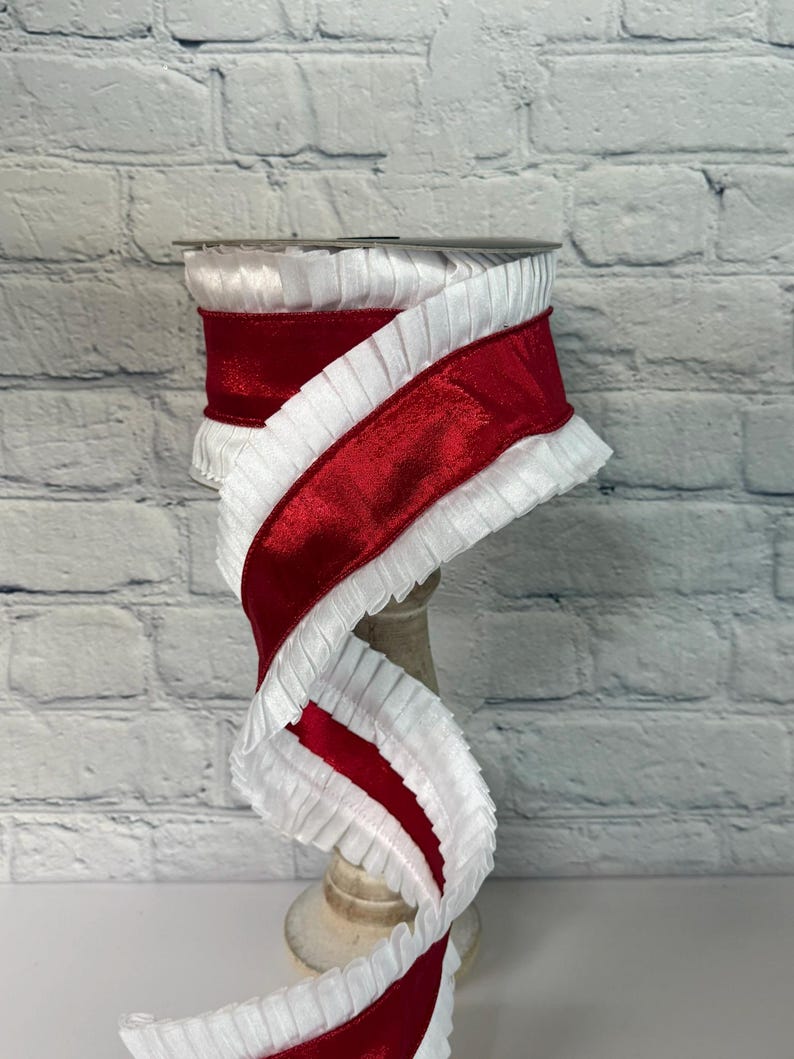 May include: A spool of red and white decorative ribbon. The ribbon features a wide, shiny red satin stripe bordered by a ruffled white fabric. The ribbon is displayed on a wooden spool, against a white brick wall.