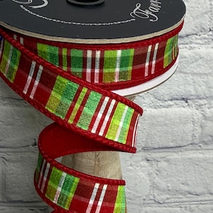 May include: A spool of wired ribbon featuring a plaid pattern in red, lime green, and white with thin metallic silver stripes. The ribbon has solid red velvet-textured edges and is wound around a black circular spool with white text.