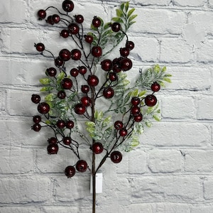 May include: A decorative artificial berry stem with dark red berries and green leaves dusted with white. The stem is brown and has a tag attached. The background is a white brick wall.