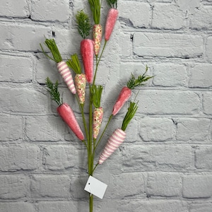 May include: A decorative Easter carrot stem with multiple carrots in varying shades of pink and patterned designs. Some carrots have white and pink stripes, while others feature floral patterns. Green faux foliage tops each carrot.