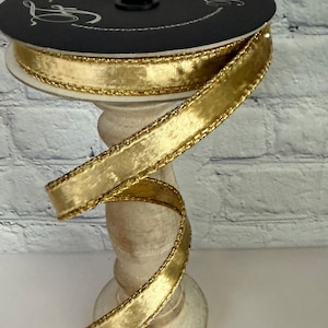 May include: A spool of gold ribbon with a metallic sheen, edged with a delicate gold trim. The ribbon is displayed on a decorative, light-colored stand, with a black label on top. The ribbon is partially unrolled, showing its texture.