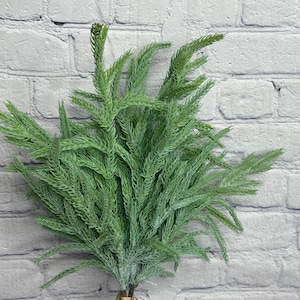 May include: A bundle of artificial green fern-like stems, with a light dusting of white, arranged in a clear glass vase. The stems are tied together with a brown twine. The background is a white brick wall.