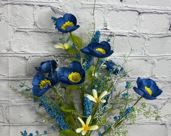 May include: A bouquet of artificial flowers featuring vibrant blue poppies with yellow centers, small white flowers, and blue bead-like accents. The arrangement is in a clear glass vase, set against a white brick wall.