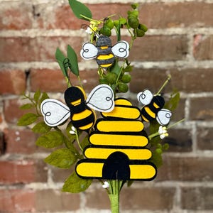 May include: A yellow and black beehive with three bees, one on each side and one on top. The beehive is surrounded by green leaves and white flowers. The beehive is on a green stem.