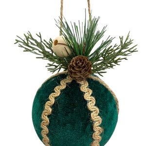 May include: A green velvet Christmas ornament with a brown pine cone and a small wooden bell. The ornament is decorated with a brown jute rope trim and has a twine hanger.