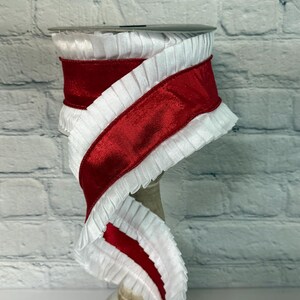 May include: A spool of decorative ribbon featuring a vibrant red velvet center bordered by ruffled white fabric. The ribbon is displayed on a neutral-toned spool, against a white brick background. This ribbon is suitable for crafts and gift wrapping.