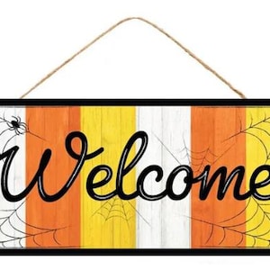 May include: A rectangular wooden sign with a black frame and a rope hanger. The sign features the word "Welcome" in black script over a background of vertical stripes in white, orange, and yellow, with spiderwebs and spiders.