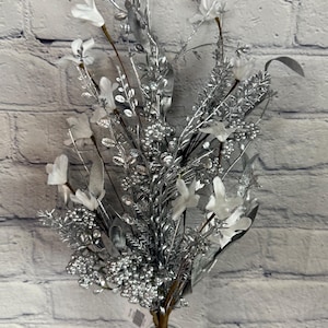 K&ouml;nnte beinhalten: Ein silber-wei&szlig;es Blumenarrangement in einer durchsichtigen Glasvase. Das Arrangement zeigt silberfarbene Zweige, Bl&auml;tter und Beeren mit wei&szlig;en Blumenakzenten. Das Bouquet steht vor einer wei&szlig;en Backsteinmauer.