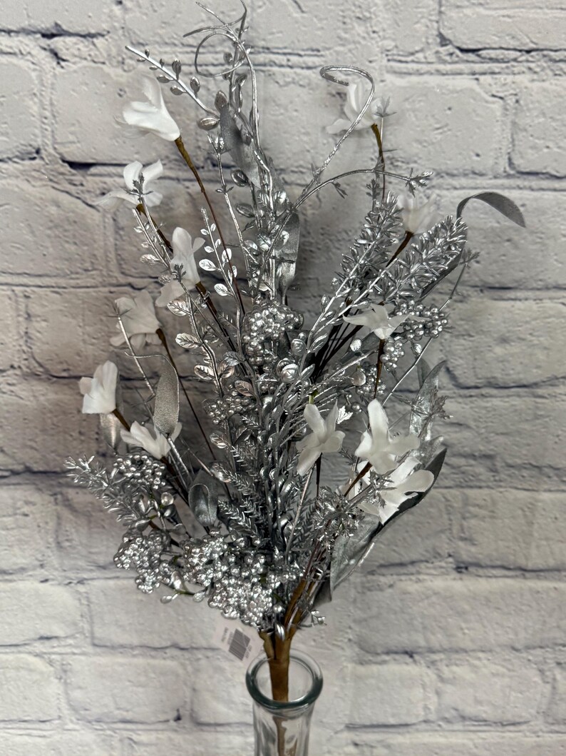 K&ouml;nnte beinhalten: Ein silber-wei&szlig;es Blumenarrangement in einer durchsichtigen Glasvase. Das Arrangement zeigt silberfarbene Zweige, Bl&auml;tter und Beeren mit wei&szlig;en Blumenakzenten. Das Bouquet steht vor einer wei&szlig;en Backsteinmauer.