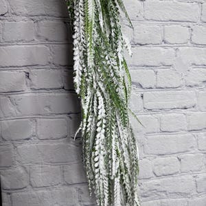 May include: A cascading artificial fern with green leaves and a dusting of white, creating a frosted appearance. The fern is against a white brick wall, showcasing its texture and color contrast. Ideal for home decor.