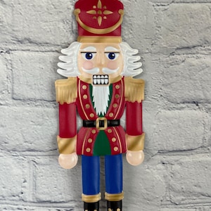 May include: A nutcracker figure wearing a red jacket with gold trim, a red hat with a gold star emblem, blue trousers, and black boots with gold laces. The figure has white hair and a white beard, with a painted face featuring blue eyes.