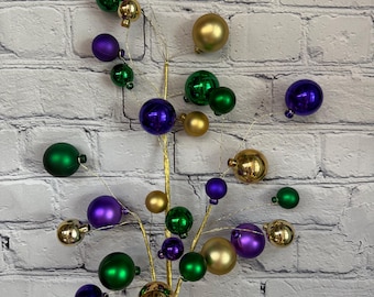 May include: A decorative spray of purple, gold, and green ornaments. The ornaments are shiny and have a metallic finish. The spray is perfect for adding a festive touch to any home decor.