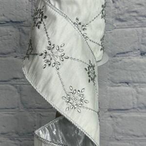 May include: A roll of white satin ribbon with silver snowflake and crystal accents. The ribbon has a wired edge and is suitable for holiday or winter-themed crafts and decorations. The snowflakes are connected by a silver beaded design.
