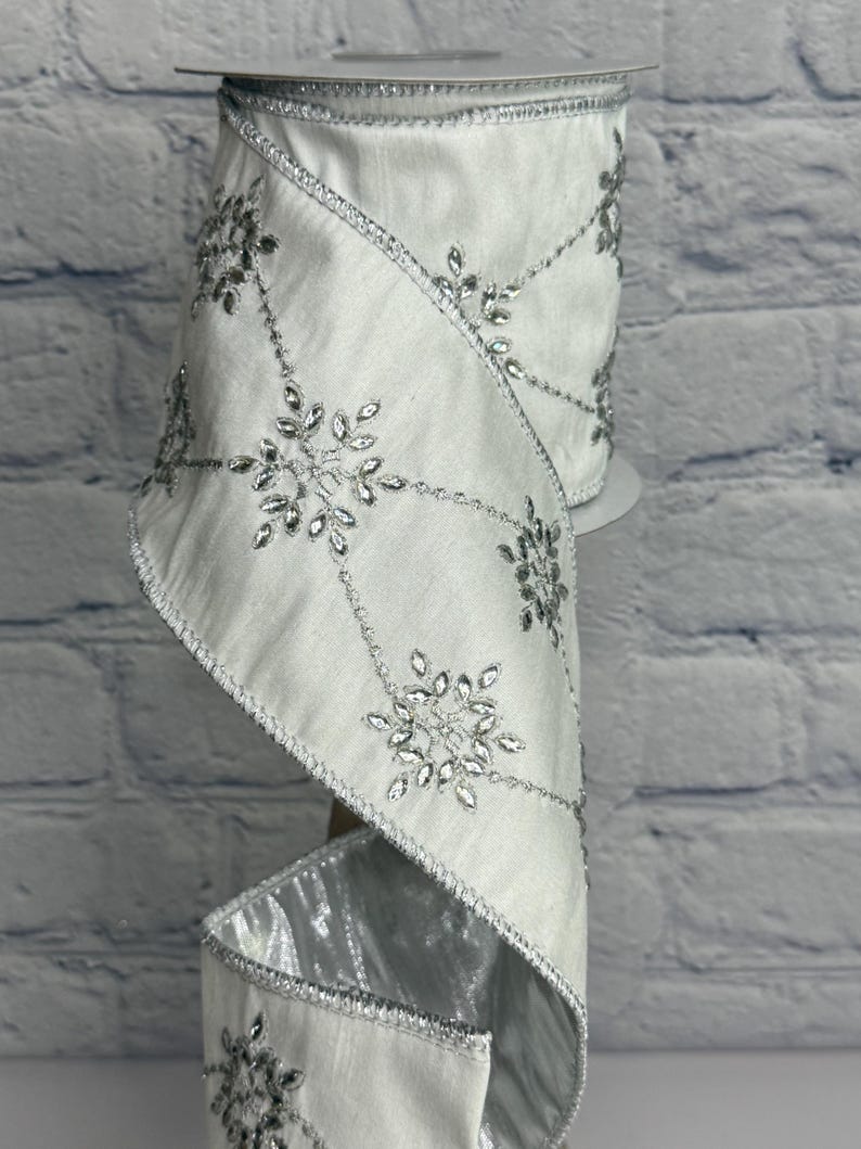 May include: A roll of white satin ribbon with silver snowflake and crystal accents. The ribbon has a wired edge and is suitable for holiday or winter-themed crafts and decorations. The snowflakes are connected by a silver beaded design.