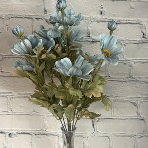 May include: A bouquet of light blue artificial flowers with green leaves in a clear glass vase. The flowers have a delicate, vintage look.
