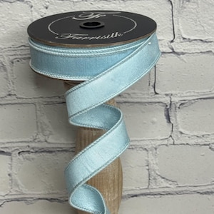 May include: A spool of light blue ribbon with a stitched edge, labeled "Farrisilk" in white script. The ribbon is displayed on a wooden spool holder against a white brick wall.