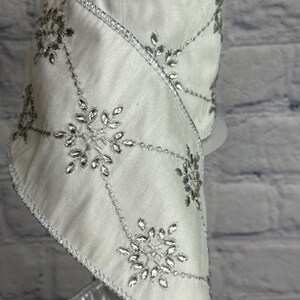 May include: White satin ribbon with silver snowflake and crystal accents. The ribbon is decorated with silver beaded lines and crystal snowflake designs. The edges are trimmed with silver metallic fabric. Perfect for holiday and winter crafts.
