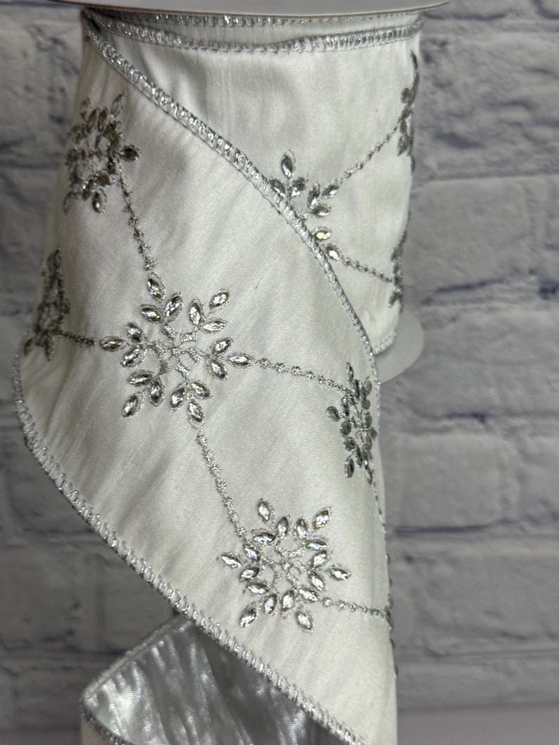 May include: White satin ribbon with silver snowflake and crystal accents. The ribbon is decorated with silver beaded lines and crystal snowflake designs. The edges are trimmed with silver metallic fabric. Perfect for holiday and winter crafts.