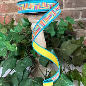 May include: A spool of decorative ribbon with a vibrant geometric pattern in shades of blue, yellow, pink, green, and orange. The ribbon is displayed on a wooden stand, with a brick background and green foliage.