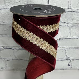 May include: A spool of deep burgundy velvet ribbon featuring a centered metallic gold embroidered leaf trim. The ribbon is wound around a black circular spool with the text Farrisilk printed in white script on the top surface of the spool.