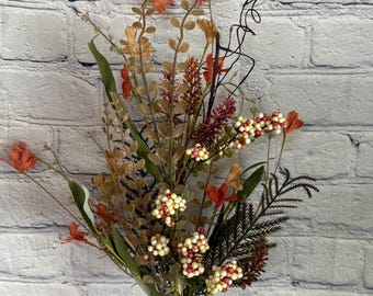 May include: A bouquet of artificial flowers with orange, red, and white accents. The bouquet features small white berries, green leaves, and brown stems.