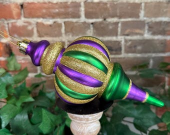 May include: A decorative Christmas ornament with a teardrop shape. The ornament features alternating stripes of gold glitter, purple, and green. It is displayed on a small wooden stand with a brick background.