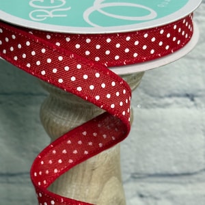 May include: A spool of red fabric ribbon featuring a repeating pattern of small white polka dots. The ribbon is wound around a circular cardboard core with a teal label that reads RG RIBBON. The fabric has a textured weave and finished edges.