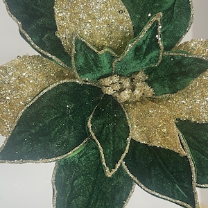 May include: Close-up of a decorative poinsettia flower. The flower features large, velvet-like green petals with gold glitter accents. The center of the flower is also gold and glittery, creating a festive holiday look.