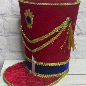 May include: A red velvet top hat with a blue velvet band at the base. The hat is decorated with gold braided trim, a gold glitter emblem featuring a dark green jewel, and a gold tassel accent attached to a small green jewel on the side.