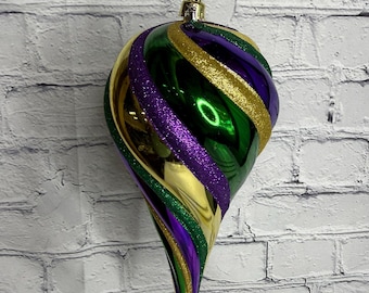 May include: A teardrop-shaped Christmas ornament featuring a spiraling design of metallic green, gold, and purple sections. The ornament includes alternating bands of glittery texture and smooth, reflective surfaces, topped with a gold cap and string.