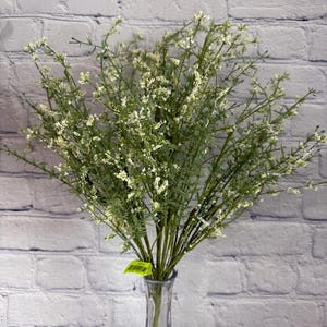 May include: A bouquet of artificial greenery with small white flowers in a clear glass vase. The stems are green, and the flowers are clustered throughout the branches. The vase is slender and sits on a white surface against a white brick wall.