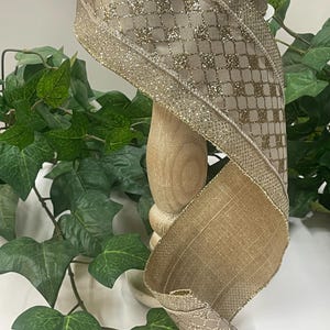 Peut inclure: Ruban d&eacute;coratif dor&eacute; et beige. Le ruban pr&eacute;sente un motif matelass&eacute; avec des accents de paillettes et une bordure dor&eacute;e scintillante. Il est enroul&eacute; autour d'un support en bois, avec un feuillage vert en arri&egrave;re-plan.