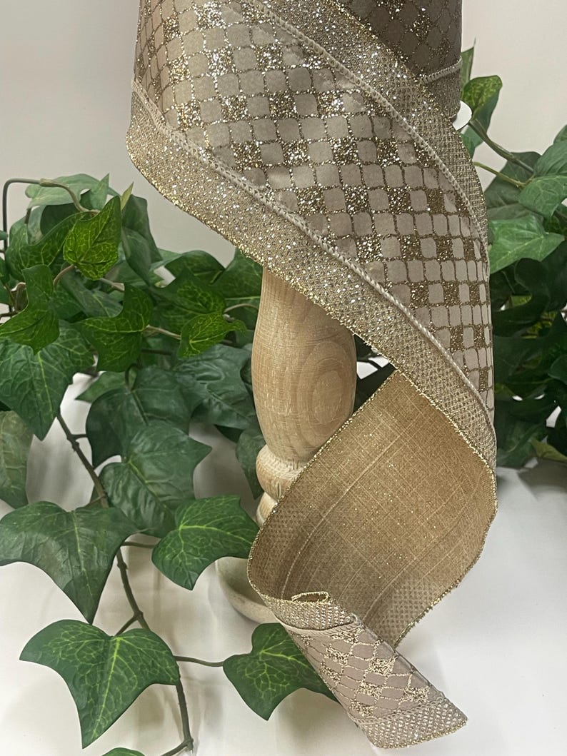 Peut inclure: Ruban d&eacute;coratif dor&eacute; et beige. Le ruban pr&eacute;sente un motif matelass&eacute; avec des accents de paillettes et une bordure dor&eacute;e scintillante. Il est enroul&eacute; autour d'un support en bois, avec un feuillage vert en arri&egrave;re-plan.